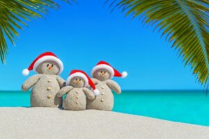 Three snowmen made from sand and wearing santa hats on a beach.
